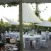 Outdoor event setup with white tablecloths, pink ribbons on chairs, and hanging lanterns under a pavilion in a garden.