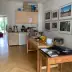 Kitchen area with wooden table, fruit bowl, cups, and framed pictures; background shows kitchen cabinets and a view into the garden.