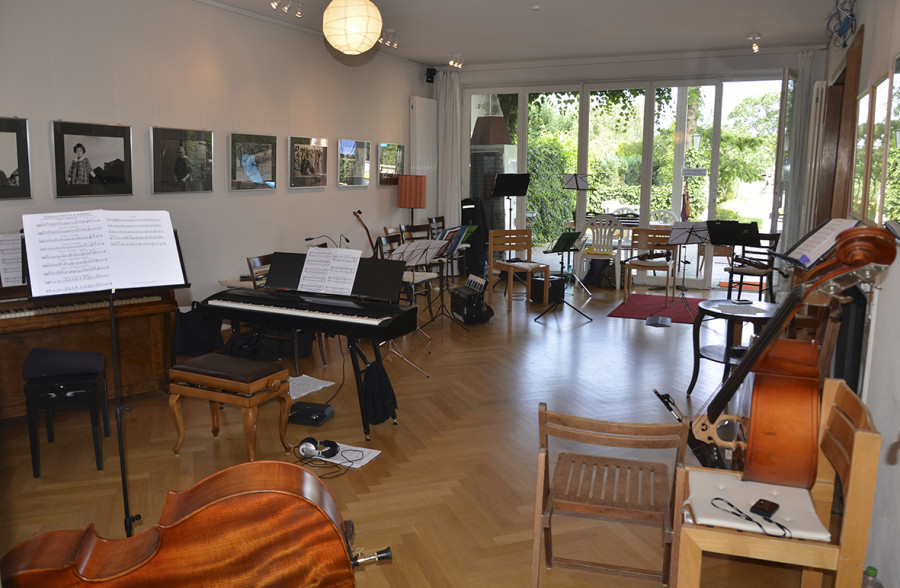Music room with piano, cello, and other instruments on stands, framed photos on walls, wooden floor, and large windows letting in natural light.