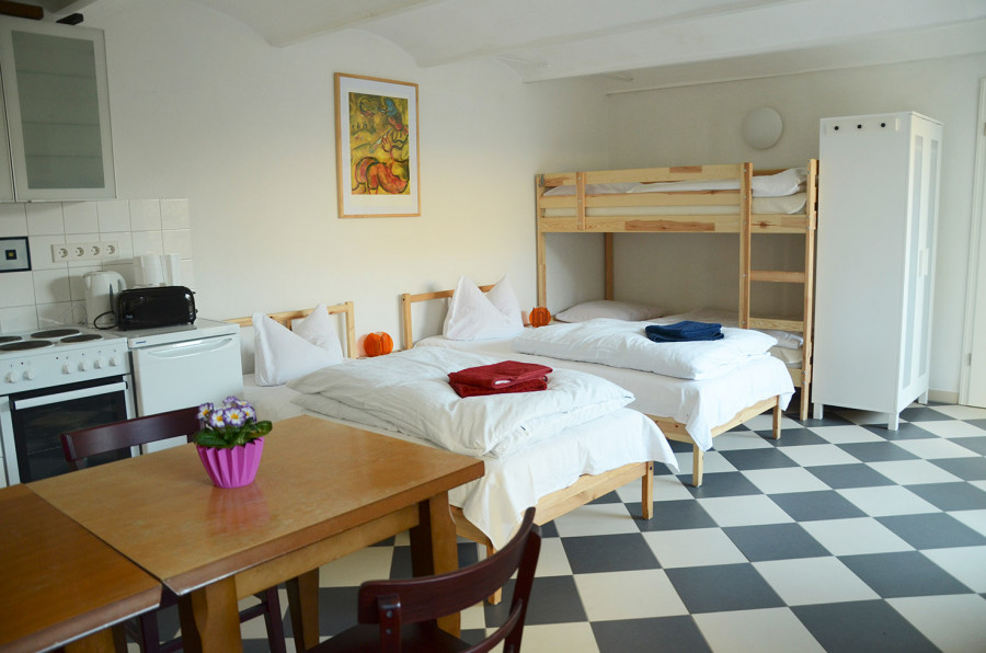 Kitchenette with stove and oven, two single beds, a double bed, wardrobe, table with chairs and flowers in room with black-and-white tiled floor.