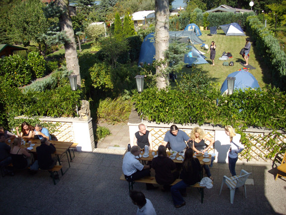 A group of people dining outdoors at wooden tables, with tents and trees visible in the background.