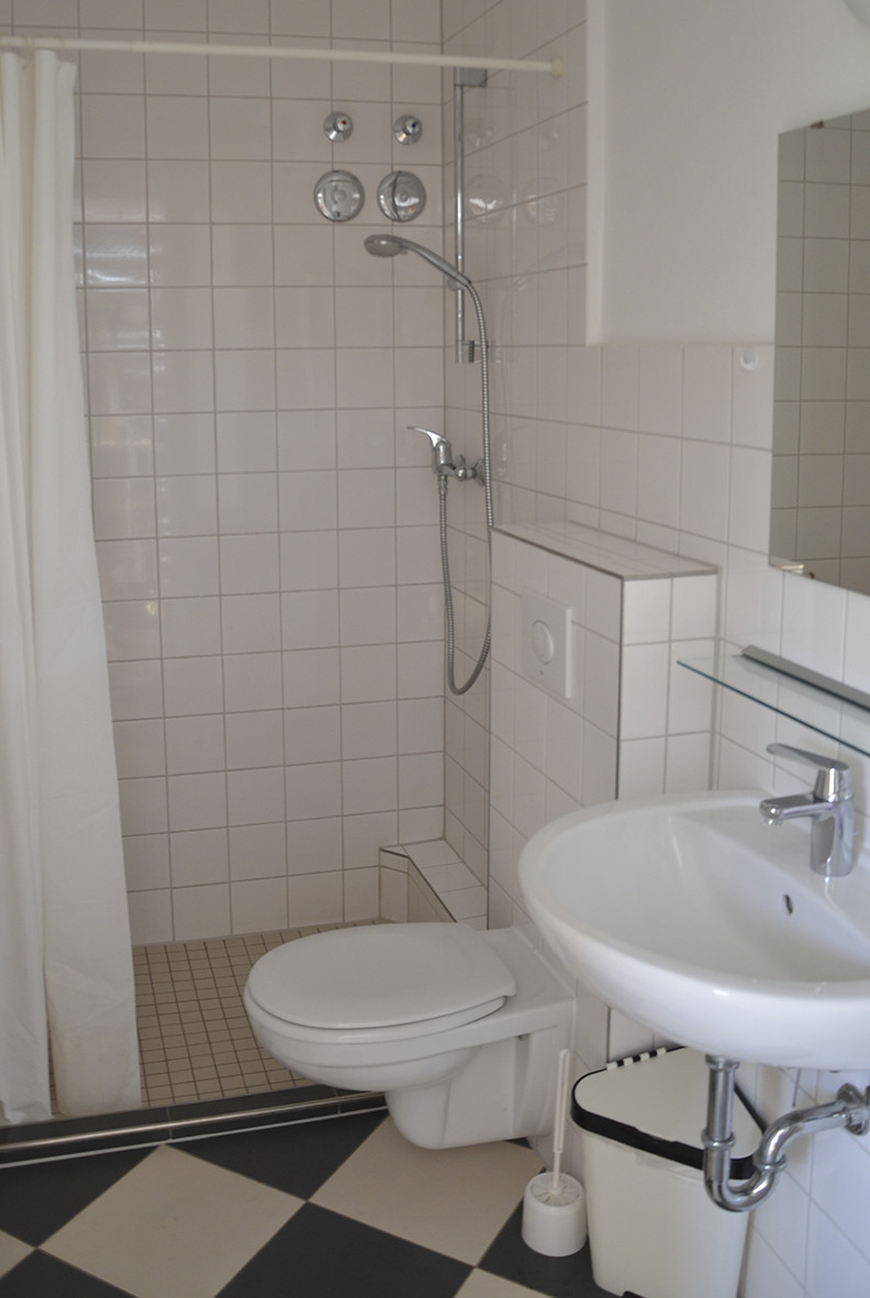 Badezimmer mit weißer Fliesenwand, Dusche, Toilette und Waschbecken auf schwarz-weißem Karomusterboden.