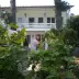 White two-story villa with balcony, surrounded by pink roses and green shrubs in a garden.