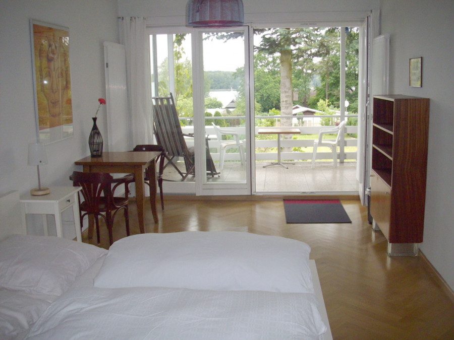 Living room with bed, wooden table, chairs and large glass window to balcony overlooking lake and trees.