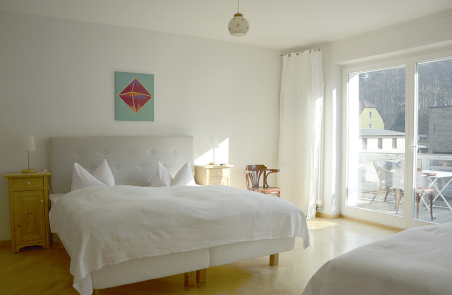 Bright bedroom with white bed, wooden nightstands, abstract painting, and view to balcony with garden furniture.