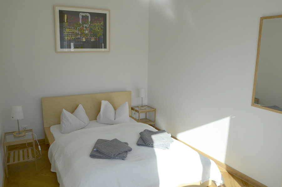 A minimalist bedroom with a wooden bed, white pillows, gray towels, and an abstract painting on the wall.