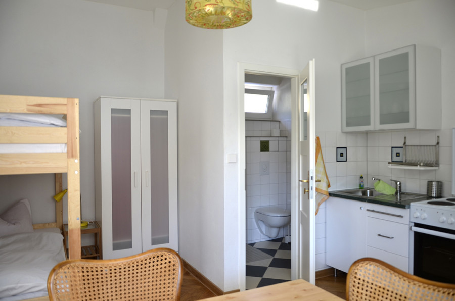 Pequeño dormitorio con litera, armario y mesa de comedor; cocina y baño visibles a través de la puerta abierta.