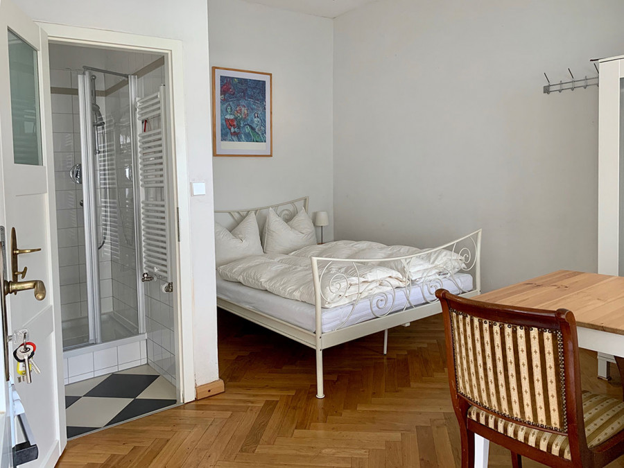 Bedroom with white metal bed, wooden floor, shower cabin, and framed wall art in a cozy living space.