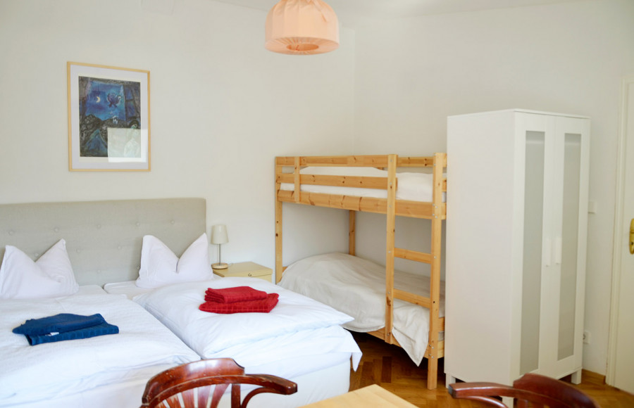 Room with double bed and bunk bed, white walls, wooden furniture, red and blue towels on beds, moon painting on wall, pink ceiling lamp.
