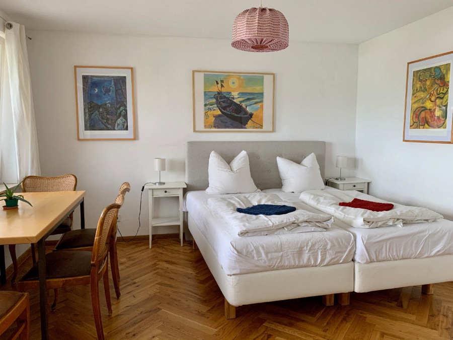 A small, brightly furnished room with a double bed, hardwood flooring, three paintings on the walls, and a dining table with chairs.