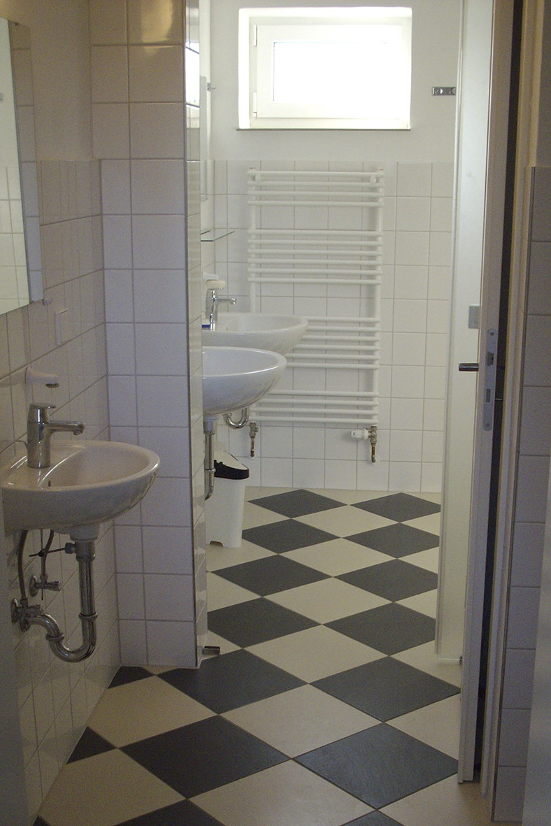 Baño con azulejos blancos, dos lavabos, radiador y suelo a cuadros negros y blancos.