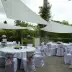 Configuraci&oacute;n de comedor al aire libre con mesas y sillas blancas decoradas con lazos rosados, bajo una gran lona blanca en un jard&iacute;n.