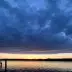 Cielo oscuro y nublado sobre un lago al atardecer con horizonte dorado y muelle de madera en primer plano.