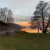 Atardecer sobre un lago con un restaurante-cafeter&iacute;a en la orilla, &aacute;rboles y una farola encendida al frente.