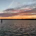 Atardecer sobre un lago con muelle, nubes anaranjadas y agua reflectante, l&iacute;nea de costa oscura al fondo