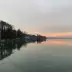 Costa de un lago con muelle, &aacute;rboles y casas al atardecer, reflejando el cielo naranja en el agua tranquila.