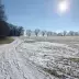 Camino de campo cubierto de nieve con rastros de neum&aacute;tico, rodeado de &aacute;rboles desnudos bajo un cielo azul y sol brillante.