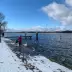 Familia con ni&ntilde;os alimentando patos y gaviotas en una orilla nevada; cielo azul con nubes, muelle de madera y &aacute;rboles desnudos en invierno.