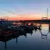 Siluetas de personas y barcos en un puerto al atardecer con cielo naranja y agua reflectante.