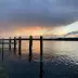 Muelle de lago al atardecer con nubes oscuras, pilotes amarillos y agua reflectante, con nen&uacute;fares en primer plano.