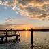 Atardecer sobre un muelle en un puerto, cielo dorado y agua reflectante, siluetas de &aacute;rboles al fondo.