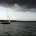 Un barco a vela con velamen blanco navega sobre aguas tranquilas bajo un cielo amenazador y oscuro con nubes, con un bosque denso al fondo.