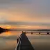 Siluetas de un muelle de madera que se extiende hacia un lago tranquilo, mientras el sol se pone detr&aacute;s de un horizonte nublado y proyecta reflejos dorados en el agua.