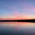 Cielo al atardecer sobre un lago tranquilo con nubes rosadas y anaranjadas que se reflejan en el agua, y &aacute;rboles oscuros al horizonte.