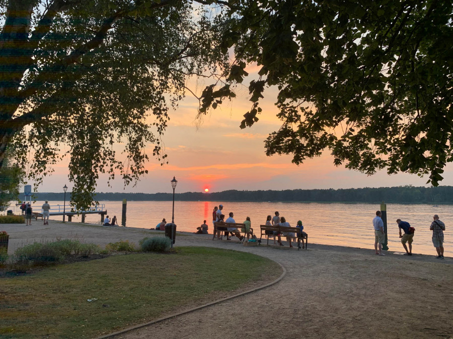 Atardecer en un sendero junto al lago, con personas sentadas en bancos y árboles que enmarcan el cielo colorido.