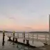 Muelle de acero con postes amarillos en la orilla del lago, horizonte boscoso y cielo rosa al atardecer.