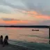 Dos ni&ntilde;os sentados en el muelle observan un atardecer vibrante de color naranja sobre un lago, mientras un barco con personas navega en el agua.