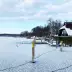 Lago congelado cubierto de nieve, muelle y caba&ntilde;a de madera tradicional en invierno, &aacute;rboles al fondo bajo un cielo nublado.