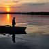 Silueta de un hombre en un bote sobre un lago al atardecer, sosteniendo una ca&ntilde;a de pescar, con reflejos dorados en el agua y un horizonte boscoso.