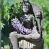 Estatua de piedra de una diosa sentada con flores moradas en sus manos, rodeada de plantas verdes al aire libre.