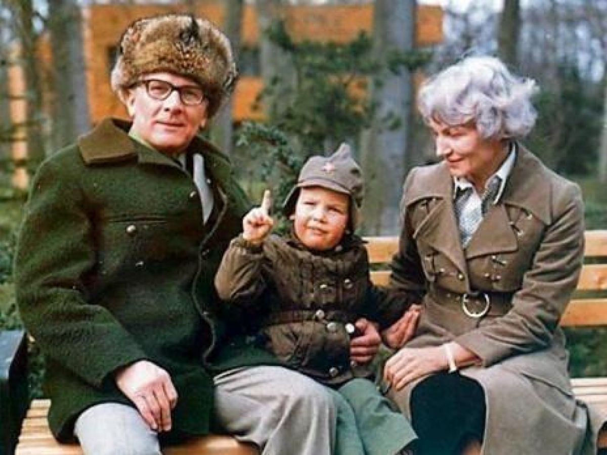 Ein Mann und eine Frau sitzen auf einer Holzbank mit einem kleinen Kind, das einen militärischen Hut trägt und zeigt. Alle tragen Winterkleidung, im Hintergrund sind Bäume und ein Gebäude zu sehen.