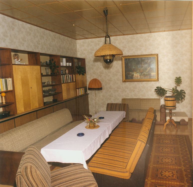 Gebäude mit Wohnzimmer, Esstisch, Sofas und Bücherregalen, dekorative Wandmalerei und Pflanzen im Raum.