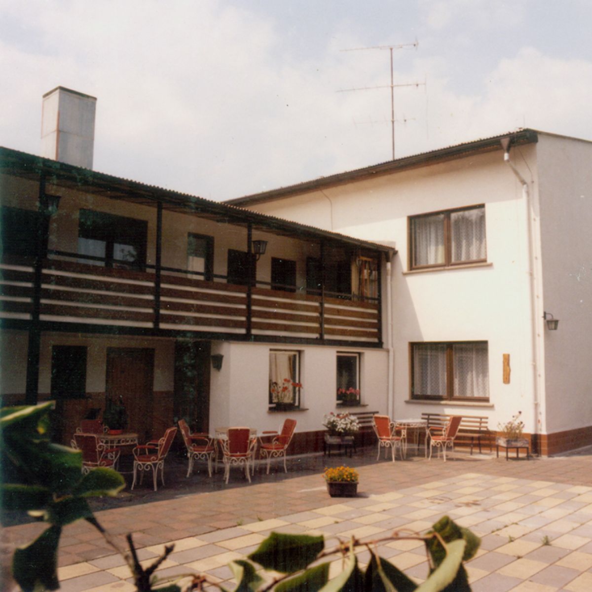 Gartenhof mit Tischen und Stühlen, Balkon, Fenster mit Vorhängen, Pflanzen in Töpfen und einem Schornstein im Hintergrund.