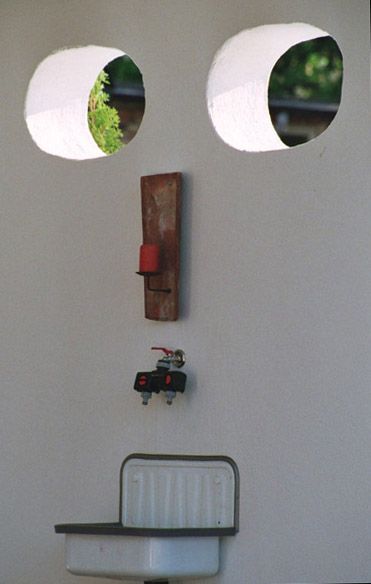 Wand mit zwei runden Öffnungen als Augen, einem Holzhalter mit rotem Kerzenständer und einem Wasserhahn über einer Waschschale.
