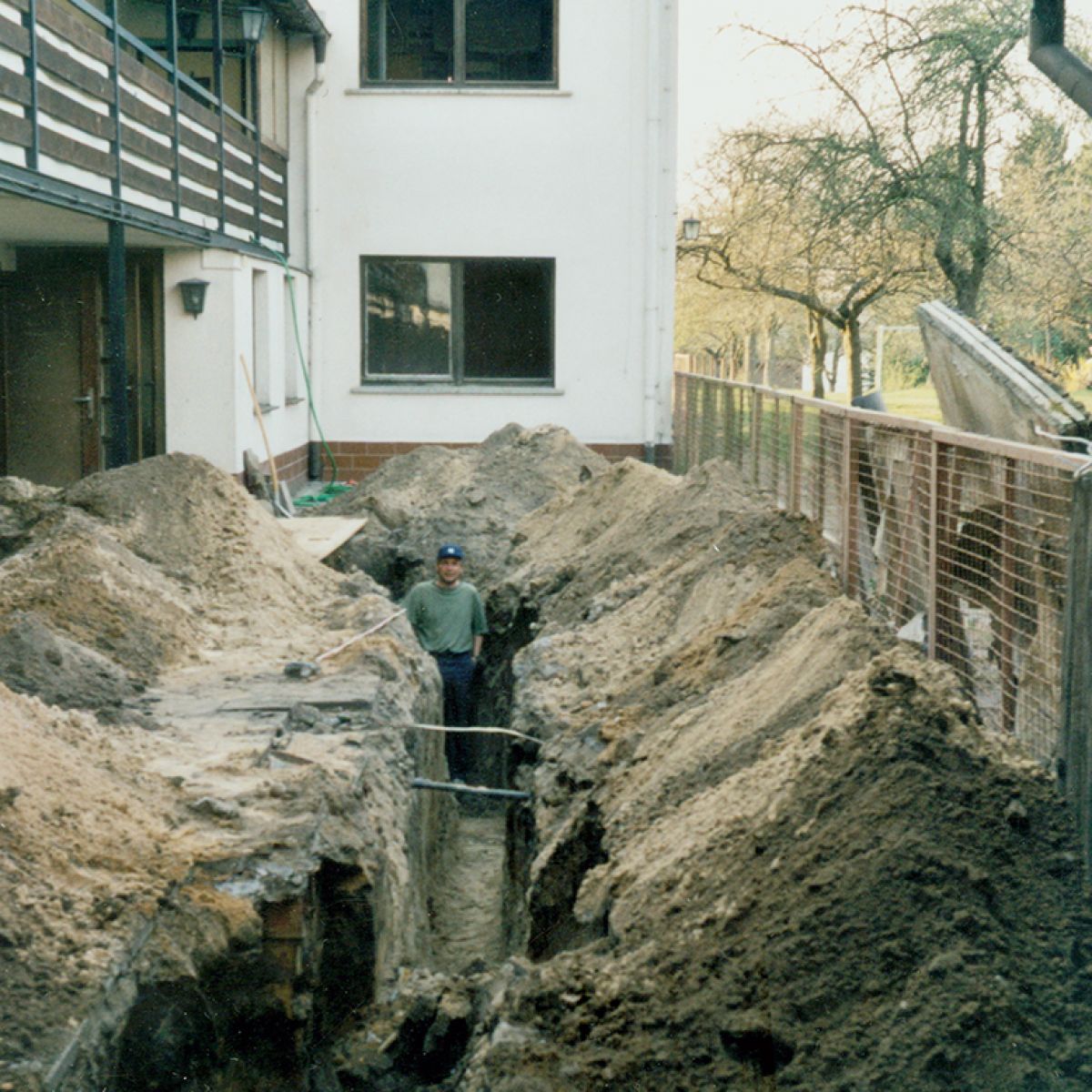 Mann in grünem T-Shirt und blauer Mütze steht in einem tiefen Graben vor einem zweistöckigen weißen Haus mit Balkon und Fenstern, umgeben von Erdhalden.
